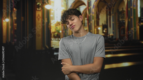 Bild auf Leinwand Teenage man in gray tee smiles with arms folded, right hand gripping left elbow, standing in ornate church building nave with pews and arches; confidence youthful ease