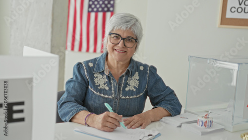 Fotografia Woman signs ballot with hand at a voting table inside a public building, american flag on wall and clear ballot box on desk; civic duty pride