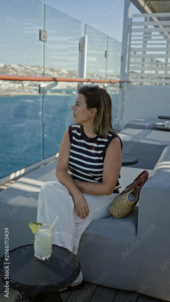 Fototapeta premium Woman sipping cocktail on cruise terrace, hand holding glass with pineapple garnish and sunglasses on head while seated on sofa; summer getaway relaxation.