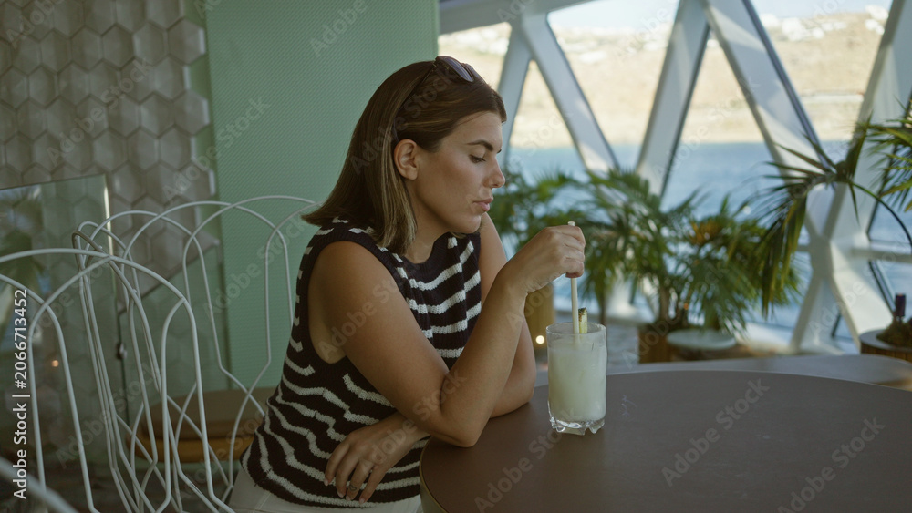 Fototapeta premium Woman stirs drink with straw at a table in a building by the water, sunglasses on head; contemplation moment.