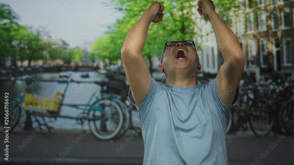 Obraz premium Man cheering with raised fists on a street beside an amsterdam canal, wearing glasses and a blue tee shirt; victory joy.