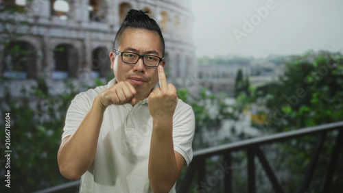 Man raising middle finger and clenched fist in front of the coliseum building while wearing glasses and white polo outdoors, facing camera; defiance travel humor.