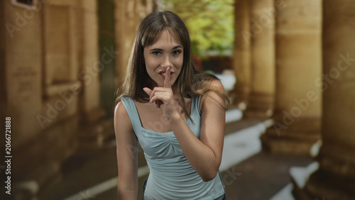 Fotografía Smiling young woman with finger to lips for silence in sunlit old town building corridor; secrecy mischief