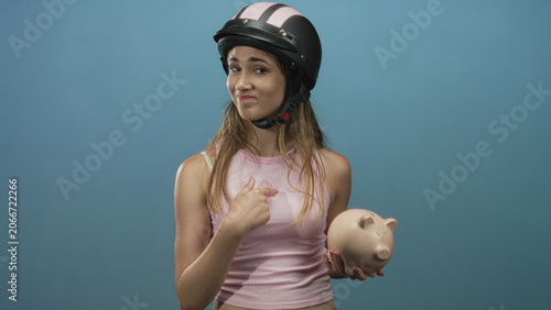 Foto Woman pointing to chest while holding piggybank and wearing helmet in studio with blue backdrop; financial doubt