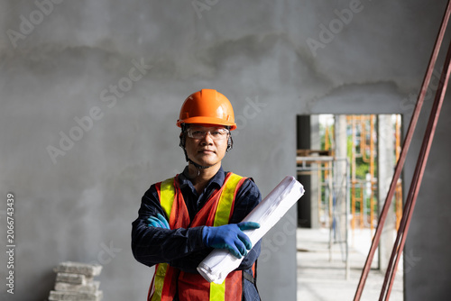 Asian construction worker in hardhat and safety vest holding rolled plans inside a building.