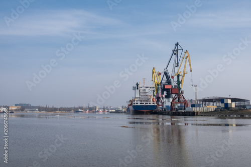 Foto Industrial harbor features a cargo ship docked next to large cranes and warehous