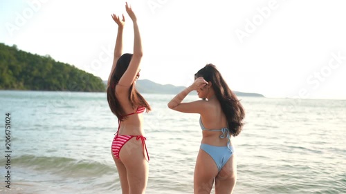 Two attractive young women in bikinis walking and laughing together on a sandy beach by the sea, enjoying a fun summer vacation, friendship, and a relaxing tropical getaway.