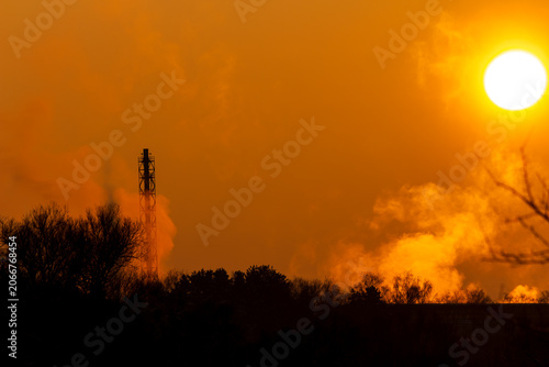 Golden winter sunset illuminates a towering industrial smokestack, its warm steam rising against the vibrant sky. Silhouetted trees below complete the dramatic scene