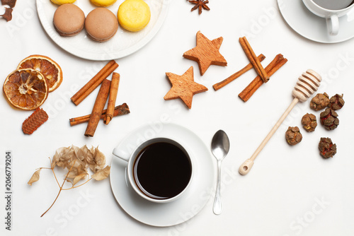 Una taza de cafe negro junto a un plato con macarons, unas ramas de canela y unas estrellas de madera decorativas sobre un fondo blanco. Vista superior y d ecerca