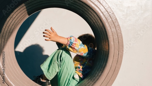 A child in colorful clothing plays inside a large circular tunnel outdoors, with sunlight casting shadows on the wall behind.