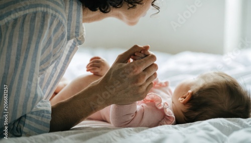 A tender moment between a parent and baby as they hold hands and smile while lying on a bed in soft natural light.
