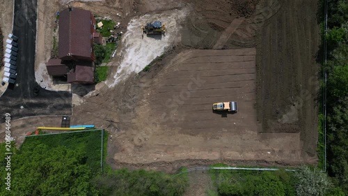 Aerial view shows a roller machine compacting and leveling the ground at a construction site near homes. Workers are preparing the area for building activities