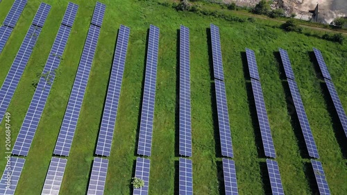 This aerial view shows rows of solar panels arranged in a large field. The panels absorb sunlight for energy. Grass and plants grow around the edges of the field