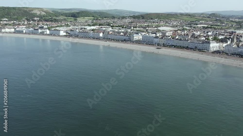 The shoreline of Llandudno is captured from above. The beach stretches along the coast with hotels lining the promenade. The water appears clear and calm on this bright day