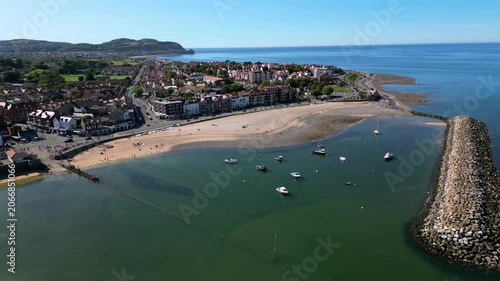 This aerial view shows Colwyn Bay Beach in North Wales during a clear day. The beach is busy with people enjoying the sun. Boats are anchored in the water near the shoreline