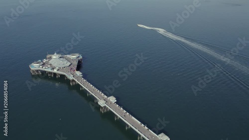 A boat travels past the pier in Llandudno. People walk along the pier and look out at the water. The sky is clear and the sea is calm