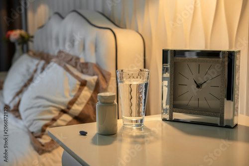 Close-up of pill bottle and water glass on bedside table with blurred bed background. Cozy bedroom interior with warm evening light, Insomnia concept and sleep hygiene