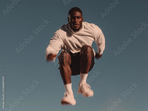 Young girl leaps mid-air against deep clear blue sky in vintage film grain texture, oversized white Nike sweatshirt paired with black shorts, dynamic tousled hair catching outdoor bright daylight.