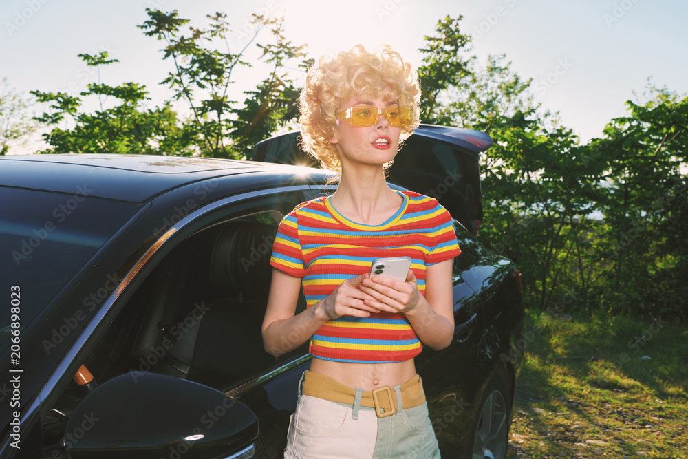 Fototapeta premium Young woman with curly blonde hair stands beside a sleek car in bright sunlight, wearing a colorful striped crop top and light jeans, enjoying a carefree outdoor moment.