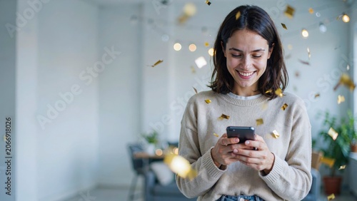 Young woman smiling while holding mobile phone during festive home celebration with gold confetti.