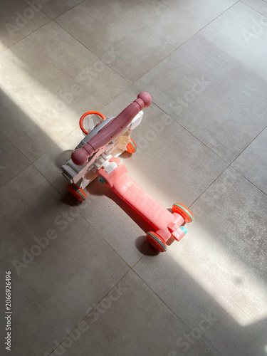 Children’s scooter resting on a polished granite tile floor as natural sunlight streams indoors, creating striking light and shadow patterns that enhance the playful and minimalist composition.