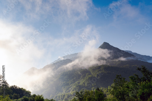 Sunlit mist drifts across forested mountain slopes near Foca, Bosnia and Herzegovina, beneath a clear blue sky.