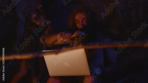 A caveman humorously using a modern laptop in a primitive setting creates an amusing contrast