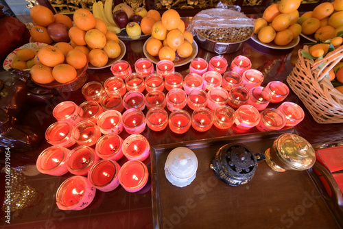 Rows of red votive candles burn on an altar surrounded by fresh fruit offerings, including oranges, bananas, and apples.