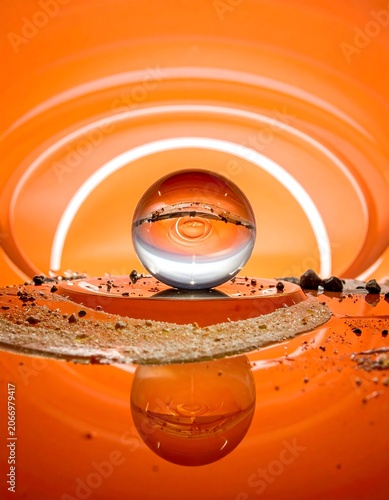 A close-up of a transparent sphere reflecting concentric circles on an orange background, with ripples in the water below