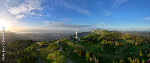 Aerial view of the Col de la Faucille mountain pass with a communication tower, winding roads, and dense forests overlooking a valley at sunset in Gex, Auvergne-Rhone-Alpes, France.