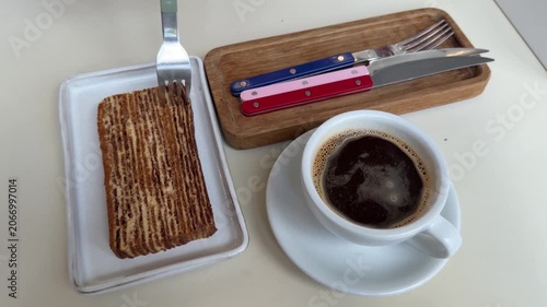 Slice of layered honey cake is cut with fork beside cup of black coffee on minimalist cafe table, dessert texture, cozy food presentation. Dessert, coffee break, hospitality, food blogging