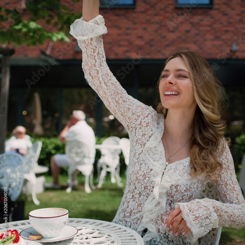 Confident woman sitting by a table at a park in an outdoor cafe with a cup of cappuccino and waving