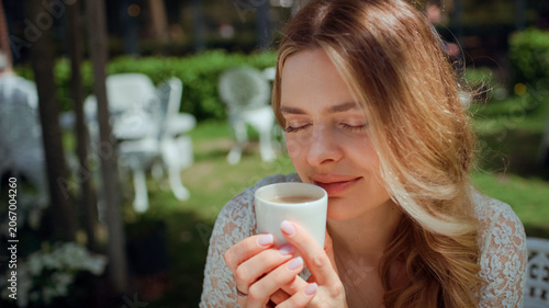 Woman sitting by a table in an outdoor cafe and holding a white cappuccino cup and enjoying the scent