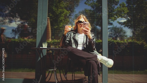 Fashionable woman sitting by a table in an outdoor cafe and holding a cappuccino while looking at her phone