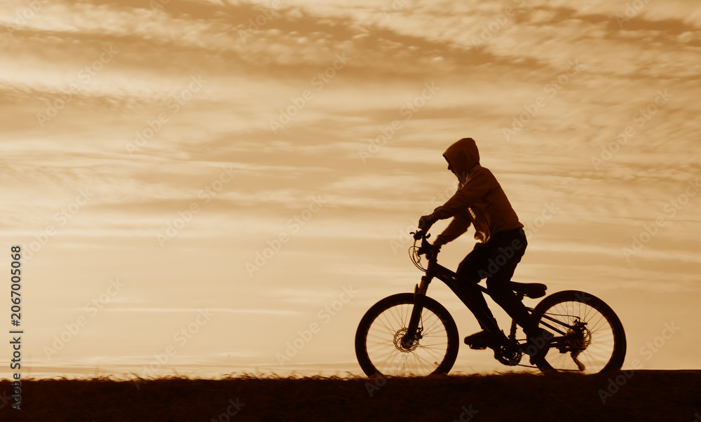 Fototapeta premium black silhouette of male young kid boy riding bicycle against yellow cloudy sky man on cycle in the nature at summer night Horizon hill line Unrecognizable person Summertime summer hot season