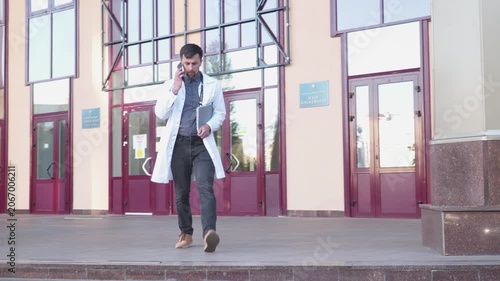 Young doctor in lab coat speak on mobile phone, leave hospital building stairs. Medical worker exiting hospital using cell phone. Medical university student walk outside building use smartphone.