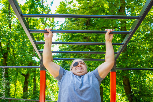 Canvas Print Middle-aged man with eyepatch doing pull-up on monkey bars in outdoor park fitne