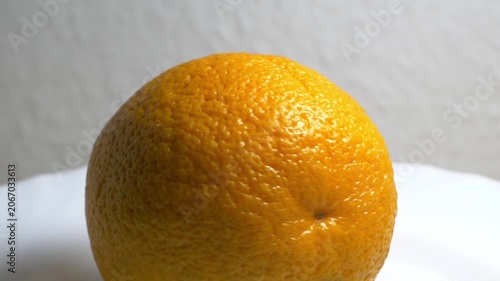 Close-up of a whole ripe orange rotating on a white turntable against a neutral background, highlighting its textured citrus peel under bright lighting.