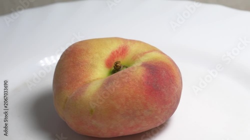Close-up of a flat donut peach rotating on a white turntable against a light background, showcasing its unique shape and fuzzy skin under studio lighting.