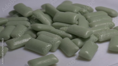 Close-up of a pile of green chewing gum pellets rotating on a white plate, showcasing their smooth glossy coating under bright studio lighting.