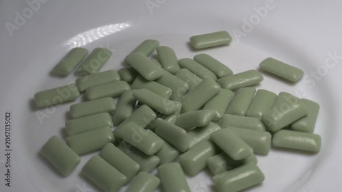 Close-up of a pile of green chewing gum pellets rotating on a white plate, showcasing their smooth glossy coating under bright studio lighting.