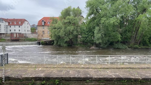 Scenic view of the Ems river weir in Rheine, Germany, with historic buildings, a church tower, and green trees lining the waterfront under an overcast sky.