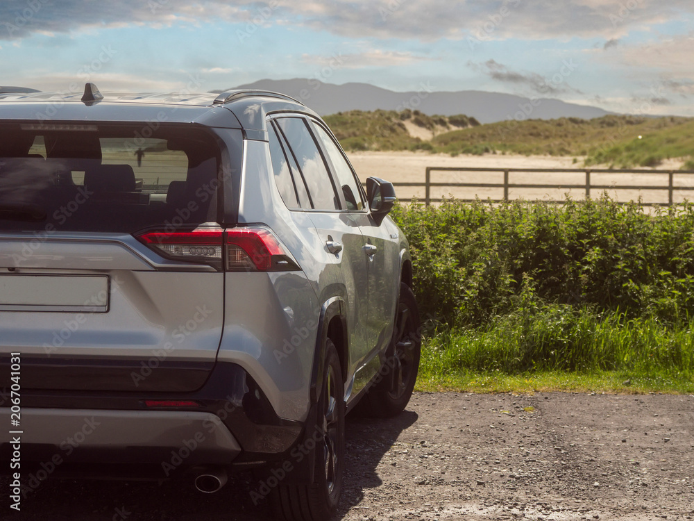 Fototapeta premium Silver SUV parked on gravel near fence. Mountainous landscape stretches in background. Green bushes line the parking lot. Sky shows scattered clouds. Outdoor travel and tourism. Rented vehicle.