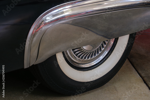 A close-up photograph of a whitewall tire and wheel on a vintage automobile. The image showcases iconic automotive styling from a classic era.