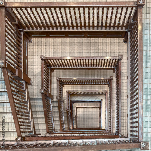 A wooden stairwell looking from the top down to the bottom. Balusters, handrails, newel posts, and carpeting are visible in the image.