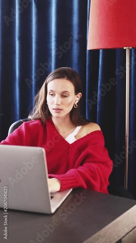 Focused Woman in Red Sweater Typing on Laptop at Desk, Taking Off Wireless Earbuds with Calm and Serious Face Expression While Working Remotely in Cozy Room with Blue Curtains and Red Floor Lamp