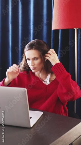 Happy Smiling Woman in Red Sweater and Earbuds Typing on Laptop, Looking at Camera with Joyful Satisfied Expression, Gathering Hair into Ponytail While Working Remotely in Cozy Room with Lamp