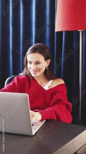 Happy Smiling Woman in Red Sweater Typing on Laptop, Looking at Screen with Joyful Satisfied Expression, Celebrating Good News or Success While Working Remotely in Cozy Room with Blue Curtains