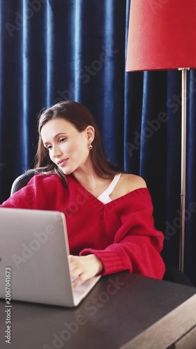 Happy Smug Woman in Red Sweater Typing on Laptop and Tilting Head with Proud Satisfied Expression, Celebrating Success While Working Remotely at Desk in Room with Blue Curtains and Lamp