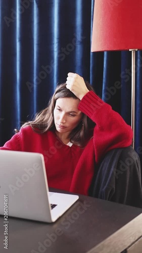 Tired and Grimacing Woman in Red Sweater Sitting at Desk with Laptop, Putting Hand on Head, Pouting and Making Funny Disappointed Face While Working Remotely in Cozy Room with Blue Curtains